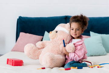 Little african american child in pink jacket playing the doctor  with teddy bear