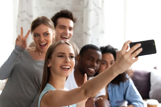 Diverse students having fun resting in cafe spends free time together, focus on female holds smartphone make selfie photo with multiracial friends smiling posing recording video for social networking