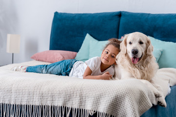 Cute little african american kid with happy dog lying on bed with colourful pillows