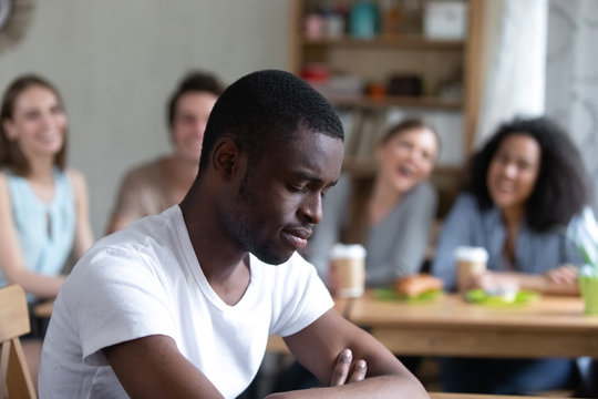 Group diverse people sitting in cafe laughing scoffing at black guy. Focus on sitting separately man feels upset and unhappy, schoolmates not take him to their company. Racial discrimination concept