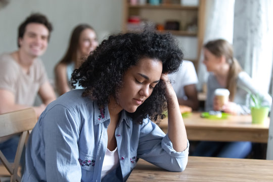 Sad mixed race girl sitting alone separately from other mates feels unhappy. Friends having fun together in cafe ignoring or scoffing at african peer. Outcast, discrimination and teen problem concept