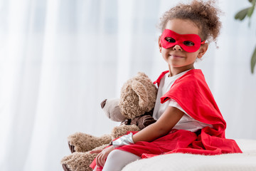Adorable little african american kid in superhero costume and mask with teddy bear