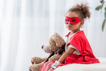 Cute african american child in red superhero costume and mask with teddy bear looking at camera © LIGHTFIELD STUDIOS