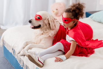 Adorable little african american kid with dog in red superhero costumes sitting on bed