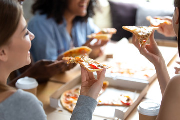 Diverse multiracial best friends students spending free time together take break have lunch sitting around table eating enjoying conversation with each other chatting, close up focus on piece of pizza