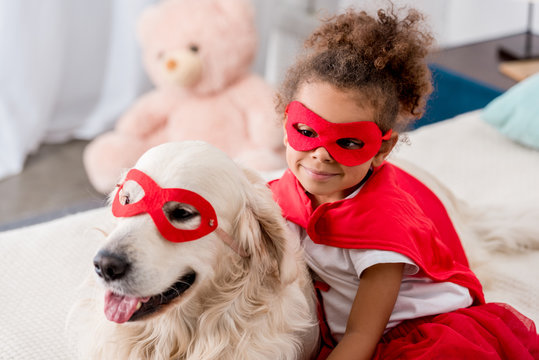 Smiling adorable little african american kid with happy dog in red superhero mask