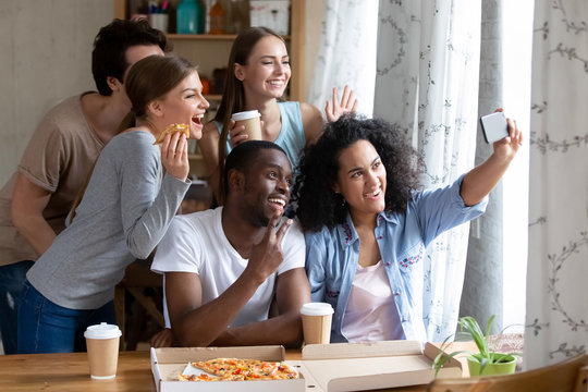 Happy diverse friends make selfie photography on smartphone during lunch. Students eating pizza and drinking coffee together indoors. Leisure activities friendship between multi-ethnic people concept