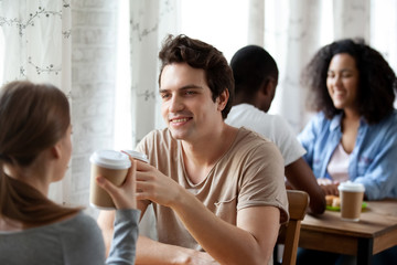 Diverse multi-ethnic couples sitting paired at table in cafe drinking hot beverages, coffee in paper cups, focus on caucasian smiling male. Friends spending time together or weekend activities concept