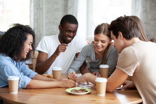 Diverse multiracial friends cheerful girls and guys drinking coffee having fun laughing assembling puzzle jigsaw sitting together at table in home or cafeteria. Teamwork and weekend activities concept