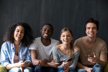 Happy diverse students multiracial friends sitting together indoor. Attractive smiling girls handsome guys holding mobile phones laughing looking at camera. Friendship and wireless technology concept
