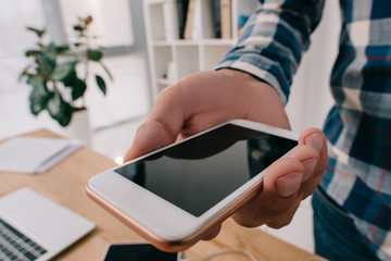 partial view of man holding smartphone with blank screen at tabletop with laptop