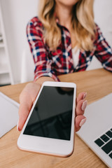 partial view of woman holding smartphone with blank screen at workplace with laptop