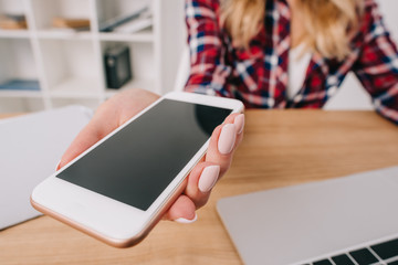 partial view of woman holding smartphone with blank screen at workplace with laptop