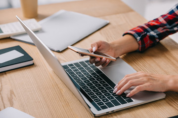 cropped shot of woman using smartphone and laptop in one time at workplace