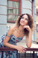 Beautiful well-groomed girl in a blue dress leaned on the railing of the old blue building. She happily smiles and dreamily looks right up.