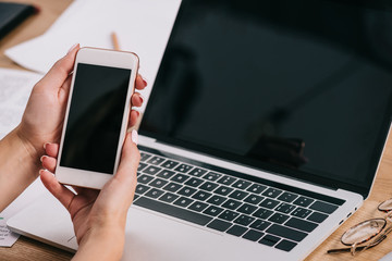 cropped shot of businesswoman holding smartphone with blank screen at workplace with laptop