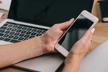 cropped shot of businesswoman holding smartphone with blank screen at workplace with laptop