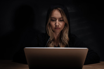 portrait of female hacker in black hoodie using laptop at wooden tabletop