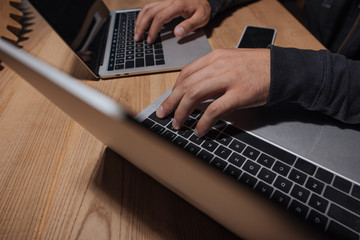 partial view of hacker using laptops at tabletop with smartphone, cyber security concept