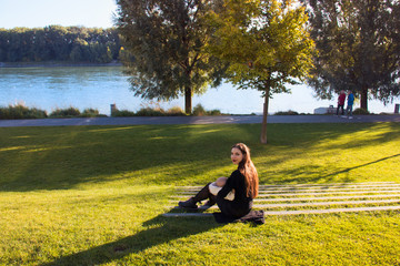 Naklejka na ściany i meble Young happy mother, white female in black breastfeeding and caring little baby sitting outside in the middle of green lawn at Danube river public promenade during wonderful autumn day
