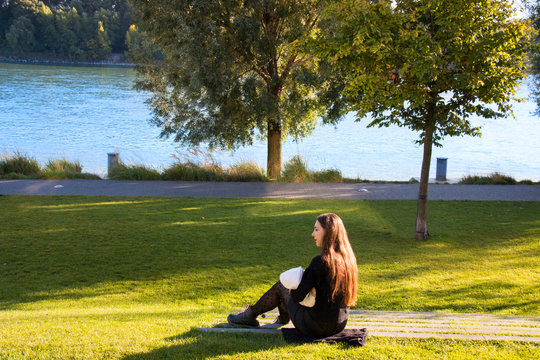 Young mother in black stickings and boots sitting on lawn outside next the stroller and breastfeeding her baby on public, autumn trees with red leaves and city building behind
