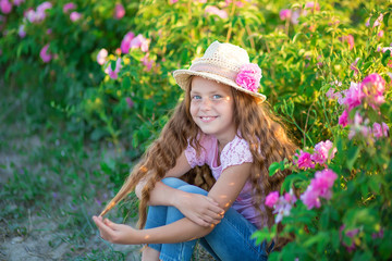 Cute brunette child blue eyes girl is holding flowers standing in pink rose field. Wearing stylish dress and hat, enjoy summer time days childhood