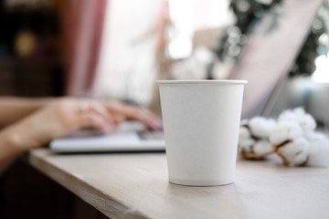 White Paper Cup of Coffee and Woman working as a Laptop on the background. Cup on workplace. Mockup. Freelance, office concept