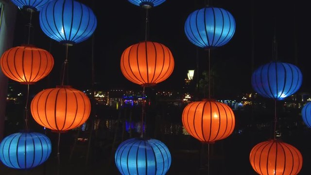 CLOSE UP: Colorful decorative lanterns illuminate the river during a historic full moon festival in Hoi An. Idyllic shot of orange and blue lamps lit up by the river during an ancient celebration.
