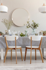 Two white chairs standing by the table with fresh plants and glass vessels in the photo of bright dining room interior with mirror on the wall