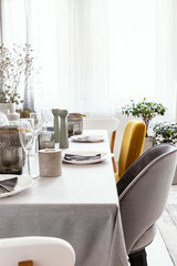 Real photo of table with dinnerware, candle and grey and yellow chair standing in bright dining room interior with window with curtains