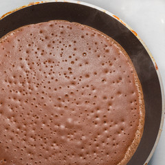 Dark chocolate pancake is fried in a pan, top view