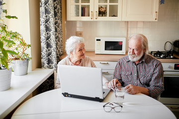 Handsome cheerful senior couple sitting at the kitchen table and using digital tablet for paying utility cost. Senior lifestyle, mature people and technology concept.
