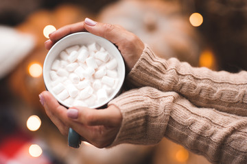 Cup of coffee with marshmallow in woman hands with Christmas lights at background.