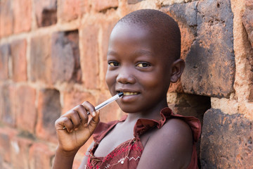 An 11-year old Ugandan girl smiling, holding a pen against her mouth and leaning against a brick...
