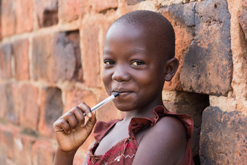 An 11-year old Ugandan girl smiling, holding a pen against her mouth and leaning against a brick...