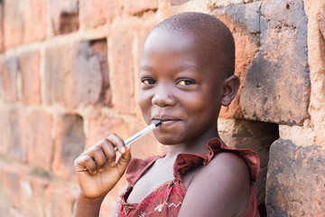 An 11-year old Ugandan girl smiling, holding a pen against her mouth and leaning against a brick wall looking at the camera