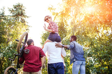 Rear view of unrecognized multiracial people going at the park in summer afternoon. Boyfriend carrying his girlfriend on piggyback and one of guys carries bmx byke over his shoulder.