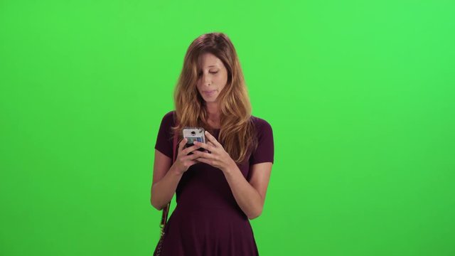 Young woman walking in a medium frontal shot over a green screen, texting happily, casual look.