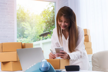 Women business owner using phone for confirm address of customer and writing in packing box at home office. online shopping SME entrepreneur or freelance working concept.