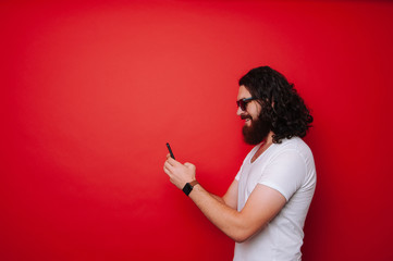 Happy cheerful bearded man with long curly hair using phone over red background