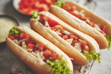 Three barbecue grilled hot dogs with sausage placed on wooden cutting board. Bowls with tomato and onionon sides. Traditional american fast food. Above angle view.