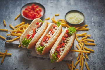 Assortment of three tasety hot dogs, placed on wooden cutting board. Served with french fries, onion and tomato. View from above. Stone background.
