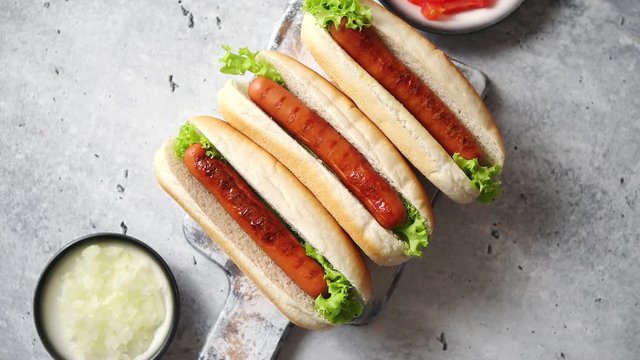 Three barbecue grilled hot dogs with sausage placed on wooden cutting board. Bowls with tomato and onionon sides. Traditional american fast food. Above angle view.