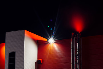 Red office building with smoke from pipe in night. Modern architecture with copy space on dark sky with lens flare. Industrial facility close up. View on below on big red wall. Shine cloud above roof.