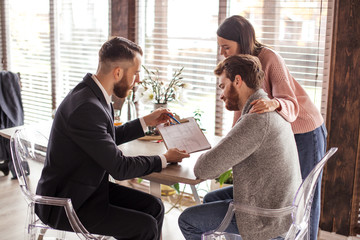 Young caucasian couple consulting with bank financial adviser before buying new house. Two bearded men sitting at table and checking documents while woman stands behind. - obrazy, fototapety, plakaty