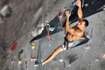 Muscular shitrless man climbing wall with belay device at an indoor wall climbing centre. Rock...