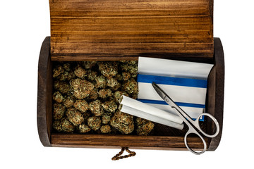 A wooden stash box half filled with green marijuana buds covered in rich red hairs with a pair of scissors, 2 prerolled joints & a pack of rolling papers on a white background.