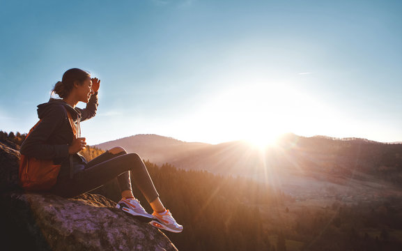woman hiker with backpack sits on edge of cliff against background of sunrise