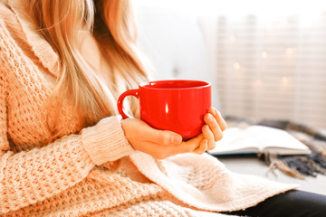 Close up young woman's hands holding big cup of hot beverage. Female wearing fashionable oversized white knitted sweater, sitting home with mug of coffee. Background, copy space.