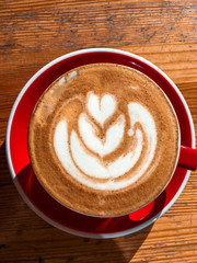 Top view of big red cup with cappuccino hot beverage with foam, with plate on wood textured table. Latte art concept. Close up, copy space, background.